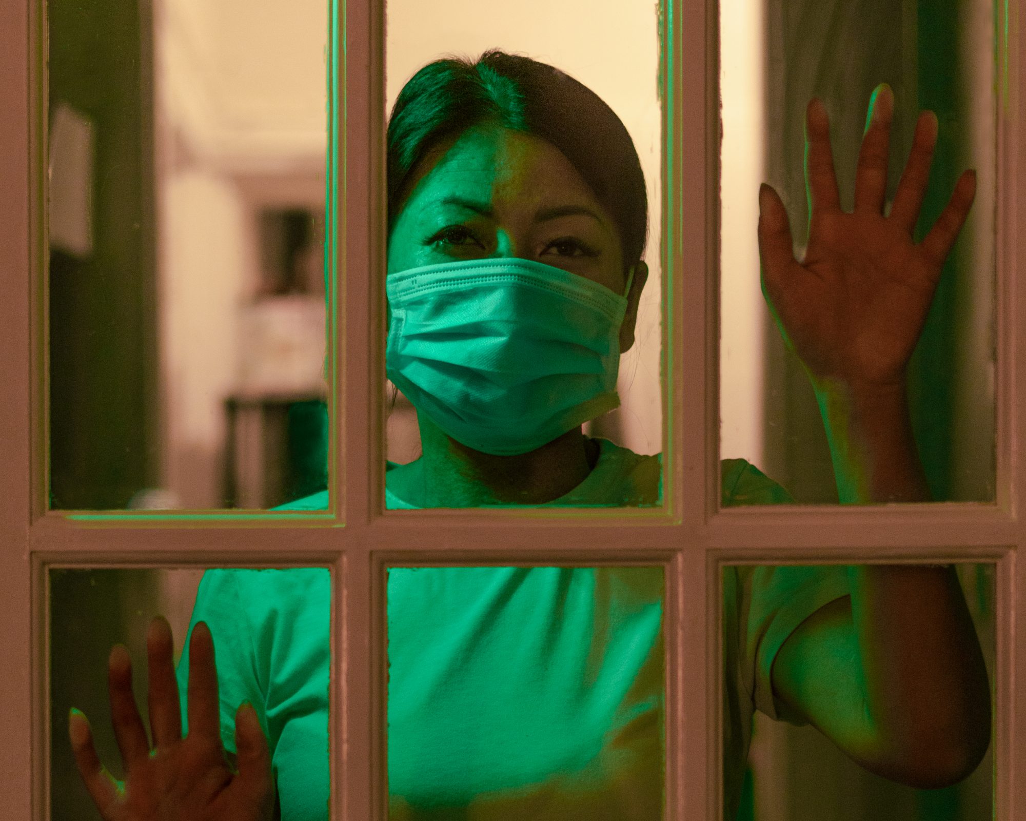 A woman looking through a window, wearing a surgical mask.